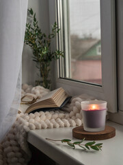 Burning candle on the windowsill by the window. Cozy windowsill with a blanket, a book, glasses. Romantic atmosphere.