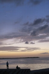 Fototapeta premium Two people sitting and standing at the pier and watching the sunset, ocean horizon idyllic scene, purple clouds in the orange sky arranging a dramatic atmosphere, perfect wallpaper to dream