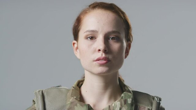 Serious young female soldier in uniform wearing body armour turning to face camera in front of plain studio background - shot in slow motion