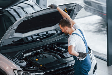 Hood is opened. Young man in white shirt and blue uniform repairs automobile