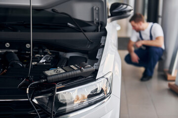 Front view of the car. Young man in white shirt and blue uniform repairs automobile