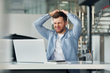 Uses laptop. Young man in white shirt and blue jacket works indoors