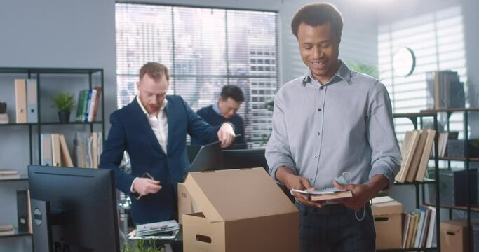 Multinational group of men have moved into new modern office and unpacking things from cardboard boxes rejoicing at their return to work.