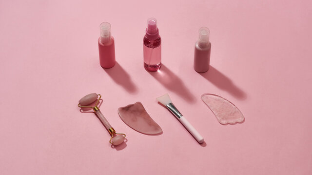 Rose quartz facial massage tools gua sha stone scrapers, face roller and beauty products in plastic bottles isolated on pink surface © Svitlana