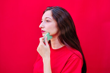 Young girl giving herself a facial massage using a gua sha stone on red background