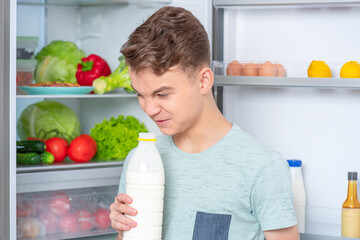 Cute young teen boy holding bottle of milk and drinks while standing near open fridge in kitchen at home. Portrait of pretty child choosing food in refrigerator full of healthy products