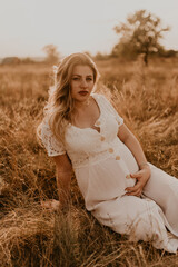 Fototapeta na wymiar Caucasian pregnant young blonde woman in cotton dress sits and lies in middle of meadow on dry grass