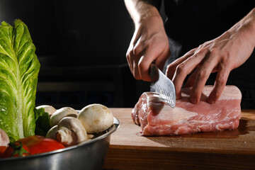 The male chef cuts raw meat with a knife on a wooden board. The restaurant kitchen cooking.