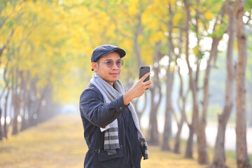 Fototapeta premium Asian man wearing sweater using mobile phone to take selfie in the park with yellow flower blossom tree during spring season