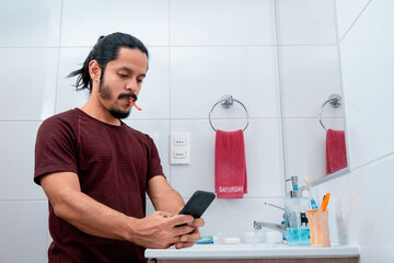Young latin man with long black hair using the phone while brushing teeth at the bathroom