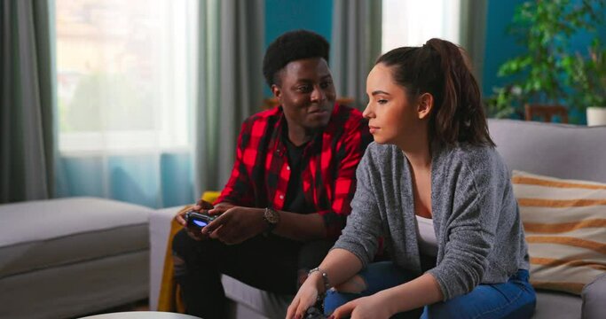 Young happy multinational couple sitting at home on couch and playing video games on console. Cheerful man having fun winning over woman in game. Boy hugs and cheers up his girlfriend