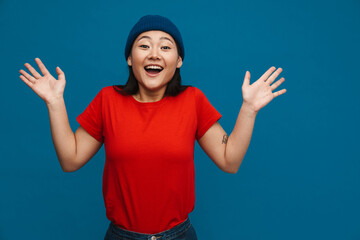 Excited asian teen girl in hat gesturing and smiling at camera