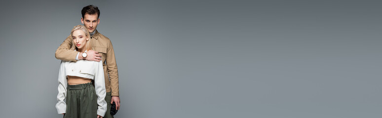 trendy man holding binoculars and hugging blonde woman isolated on grey, banner