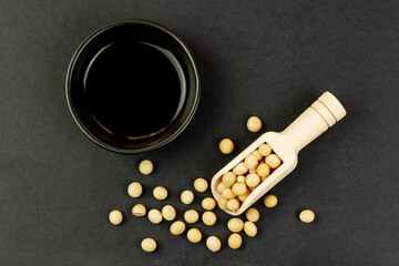 Soy sauce in small black bowl with soybeans on wooden spoon on black background, top view. Soya sauce as a cooking ingredient.