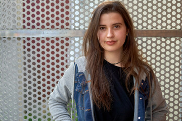 Young girl with long hair and light skin wearing a blue denim jacket leaning on a railing looking at the camera with unfocused urban background. teenager with street look in a city