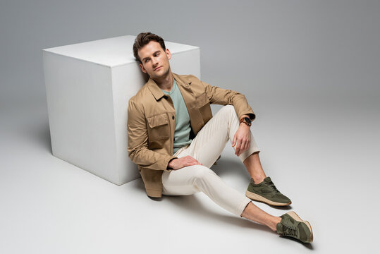 full length of stylish man posing while leaning on white cube on grey