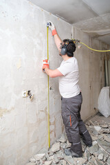 Builder man using a measuring tape at the construction site. Construction worker wearing safety gloves, glasses and earmuffs.