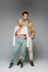 Fototapeta premium full length of trendy models in pants and jackets posing with hands in pockets on grey