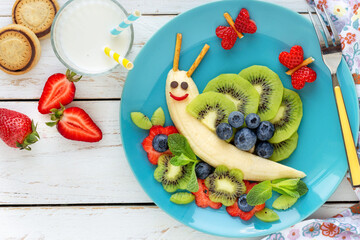 Fun food for kids - cute smiling snail made of fresh fruits (bananas, kiwi, blueberries and strawberries) as a healthy breakfast for children served with milk and cookies