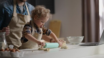 cute toddler with funny curls is playing at kitchen, rolling out dough, master-class from father, dad and son