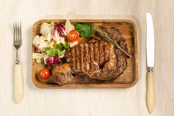 A portion of Prime beef steak on a wooden plate and a fresh tomato salad. Fork and knife cutlery on the table.
