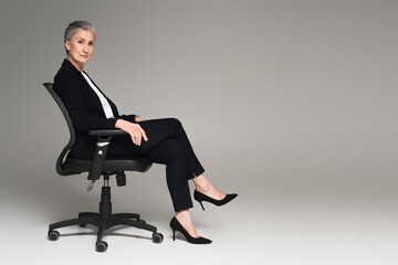 Middle aged businesswoman with crossed legs looking at camera on office chair on grey background