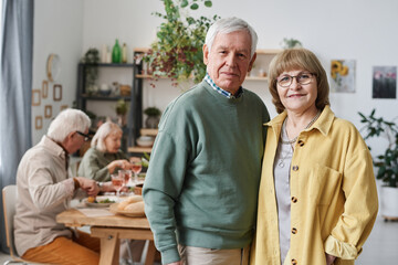 Portrait of senior couple looking at camera while standing in the room with their friends in the background