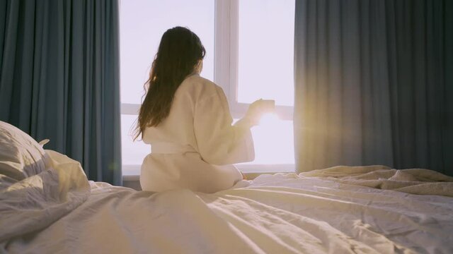 Tranquil young woman in white bathrobe drinks coffee sitting on large comfortable bed near panoramic window in hotel room at sunrise backside view