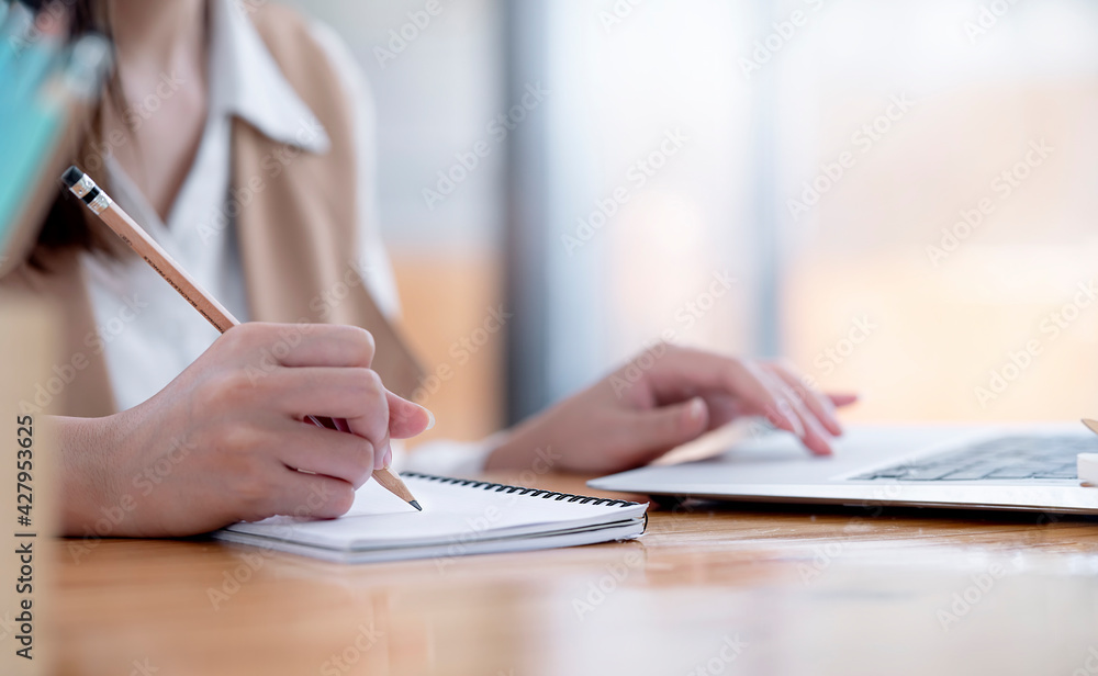 Poster closeup image of woman hands writing on notebook and using laptop computer while sitting at home off