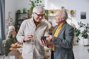 Two senior men holding glasses of red wine using mobile phone and discussing online work