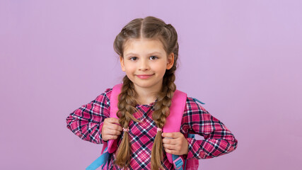 A little girl holds a school satchel and smiles against a pink isolated background.