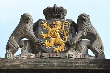 A powerful stone sculpture of the Belgian coat of arms with a crowned rampant lion in yellow at the center, flanked by two guarding lions holding shields atop a building in Bruges
