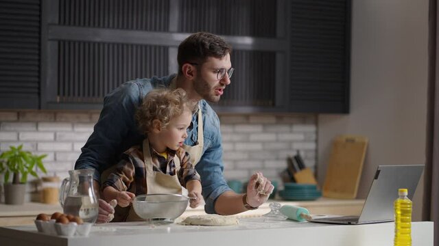 man and his baby son are cooking by internet recipe, man is reading info in laptop and toddler is playing with dough