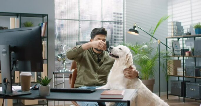Young handsome Hindu guy is sitting in office with his big dog having fun feeding him pet treats.