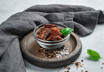 Healthy dried meat with pepper and salt is decorated with basil in a transparent cup on a wooden plate on a gray background.