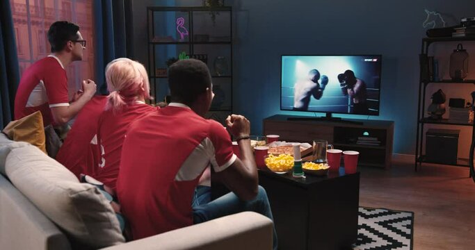 Mixed-race differente happy friends males and females sitting on couch in living room at home in evening watching sport program on TV, box fans cheering for favorite boxer. Entertainment concept