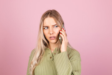 Pretty european woman in casual knitted sweater on pink background isolated hold phone having conversation with curious confused serious face