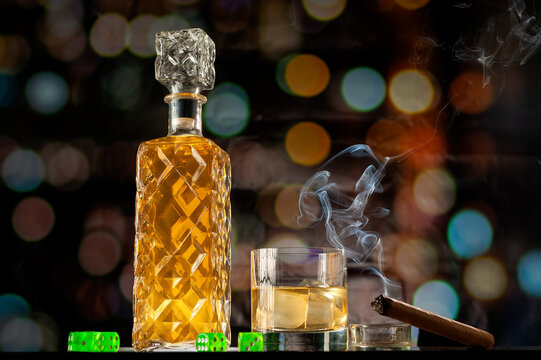 carved whiskey bottle with glass full of whiskey and ice with smoking cigar and green acyl dice with defocused lights background