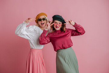 Two happy smiling playful girls, friends, sisters, wearing trendy sunglasses, colorful blouses, skirts,  berets, posing on pink background. Spring fashion, advertising concept. Copy space for text