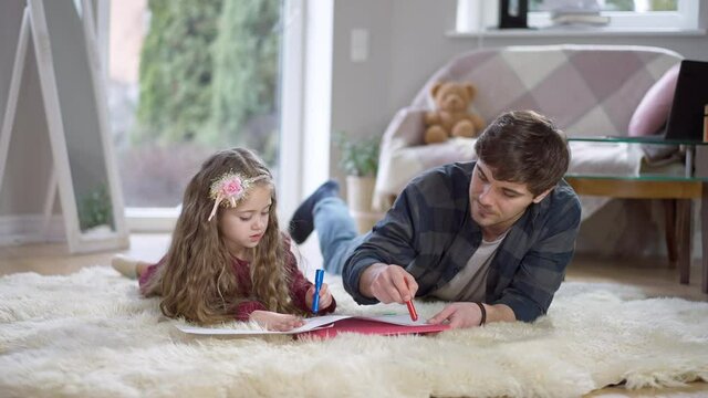 Relaxed young Caucasian father and pretty daughter lying on soft carpet drawing picture at home. Portrait of carefree parent and child enjoying weekend together indoors. Lifestyle and unity