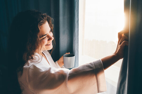 Curly haired lady in white bathrobe opens curtains to look outside large window drinking hot coffee in dark hotel room at sunrise time.