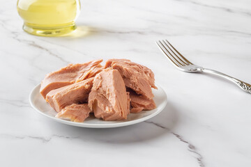 Canned tuna fillet pieces on a white saucer and fork over marble table close-up. Preserved tuna...