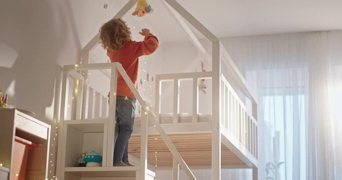 Little blond boy with curly hair happily hangs a garland in his nursery climbs the stairs of a bunk bed smiles