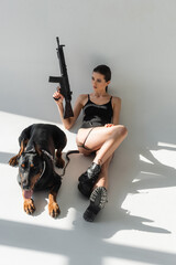high angle view of sexy woman with rifle near doberman dog on grey background with shadows