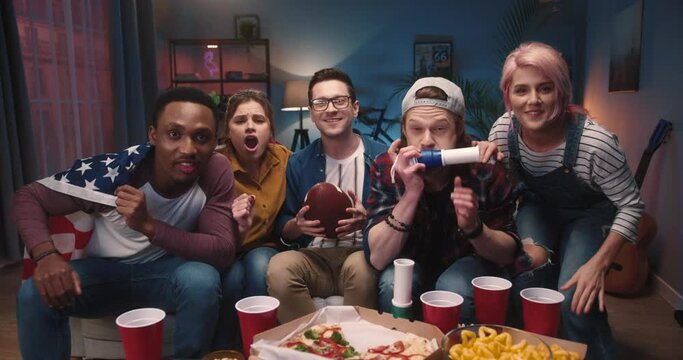 Multi-ethnic cheerful happy young people sitting on sofa in apartment at night watching television feeling excited cheering for fav soccer team at home. Entertainment, sport fans, football concept