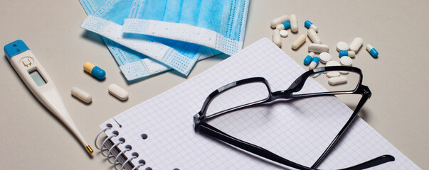 On a light gray background, a disposable face mask, an electronic thermometer, pills, a notebook and black-framed glasses. Medical concept