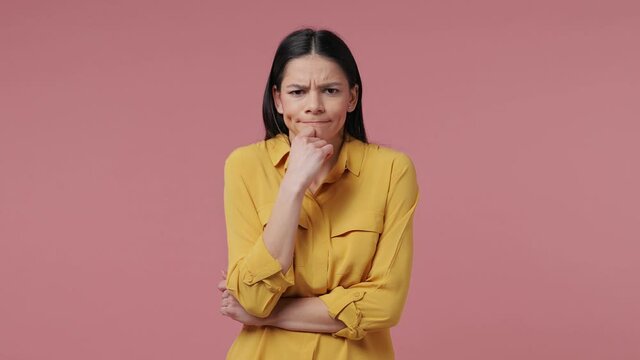 Puzzled confused brunette young latin woman 20s in casual yellow shirt posing isolated on pastel pink background studio. People lifestyle concept. Spreading arms put hand prop up on chin say oops