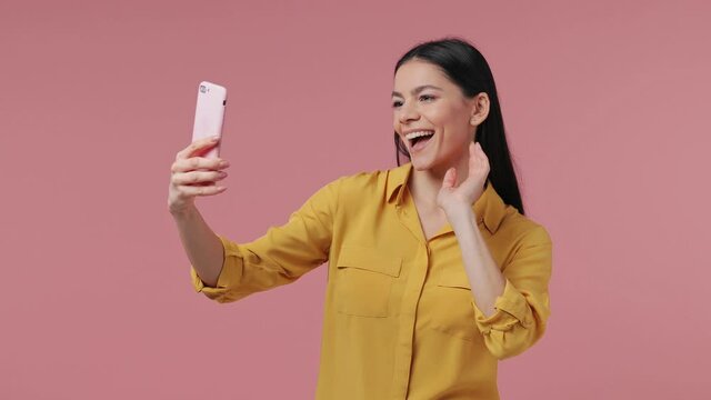 Funny young latin woman in yellow shirt posing isolated on pink background studio. People lifestyle concept. Doing selfie shot on mobile phone showing victory sign thumb up like blowing send air kiss