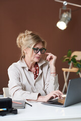 Mature businesswoman in eyeglasses sitting at the table and typing on laptop at office