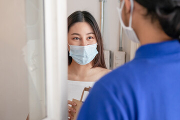 Young asian woman, customer wearing face mask protect to pandemic of covid and received bag,cardboard box from delivery man while lockdown,quarantine at entrance, door of home,house.Take away concept.